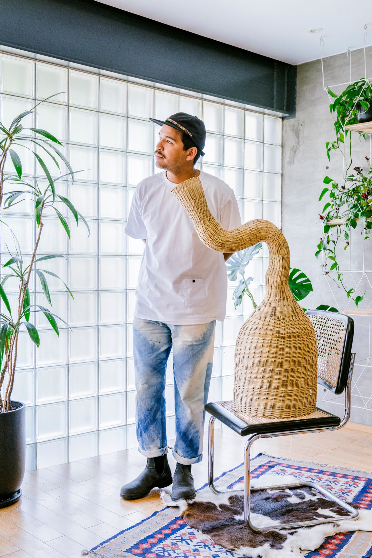 Man in white shirt and jeans standing next to woven basket sculpture in bright interior with plants