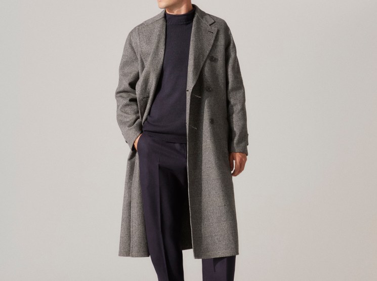Model wearing gray plaid coat over navy sweater with dark trousers and black leather shoes