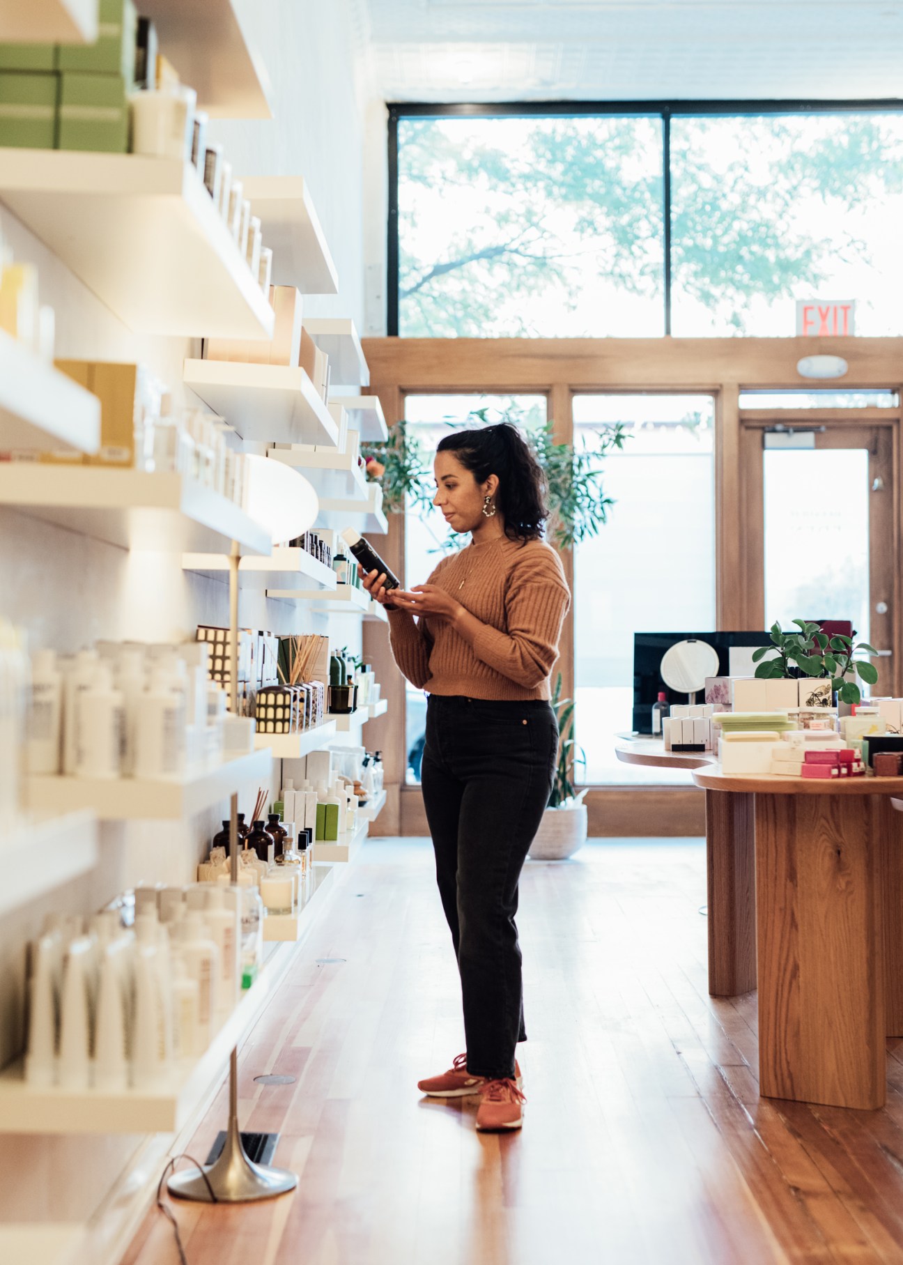 Woman browsing products on white shelves in a bright, minimalist beauty and lifestyle shop with wooden counter and large windows.