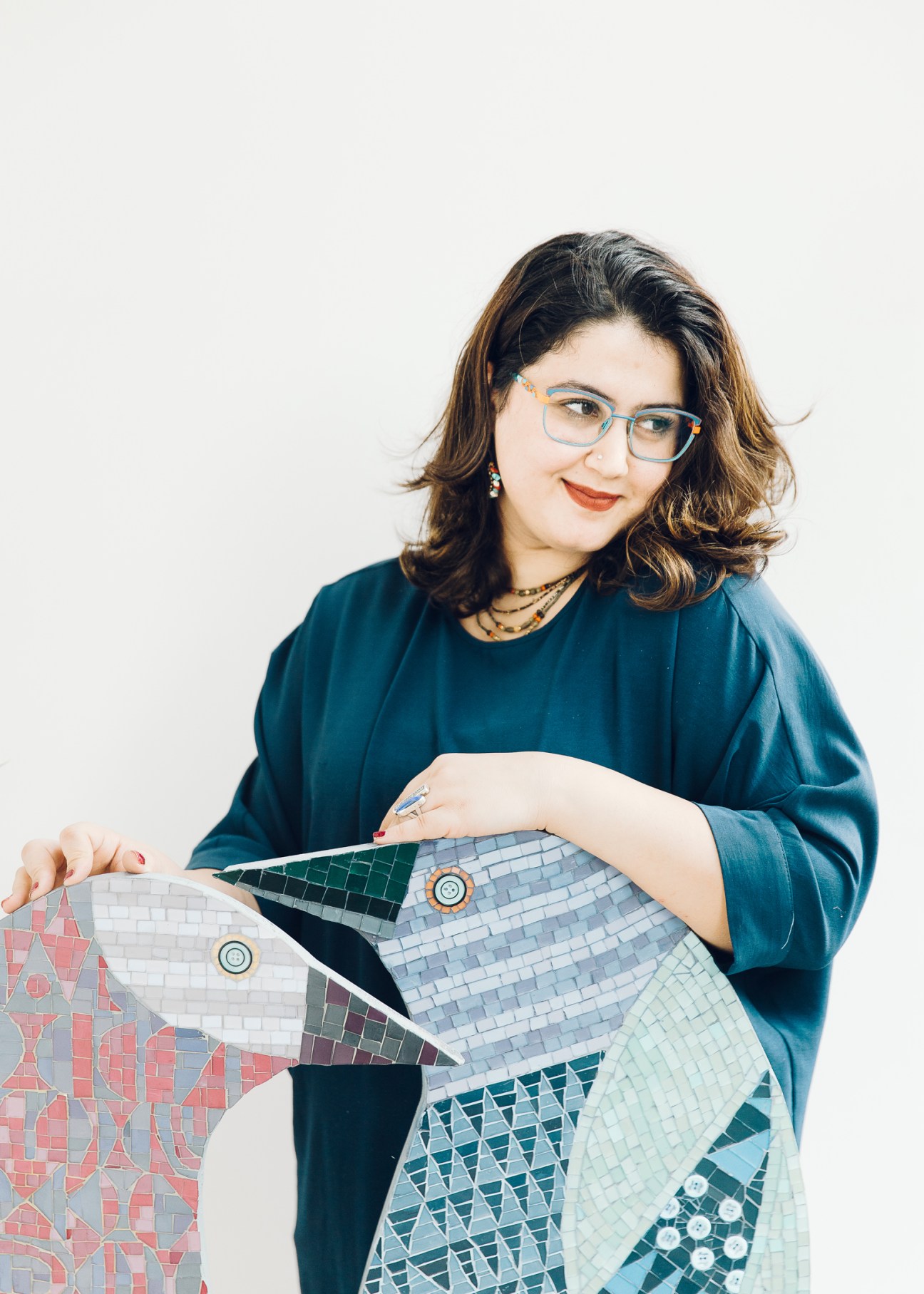 Artist Noor Ali Jaber wearing glasses and a teal top, holding colorful mosaic artwork with geometric patterns