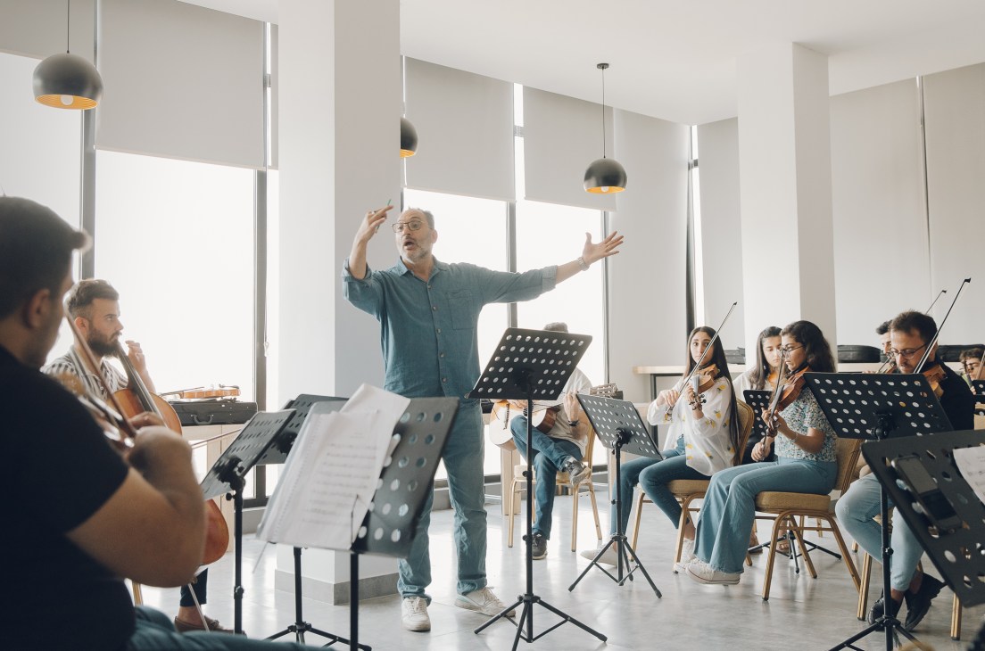Orchestra conductor with raised arms directing string musicians in a bright modern rehearsal room