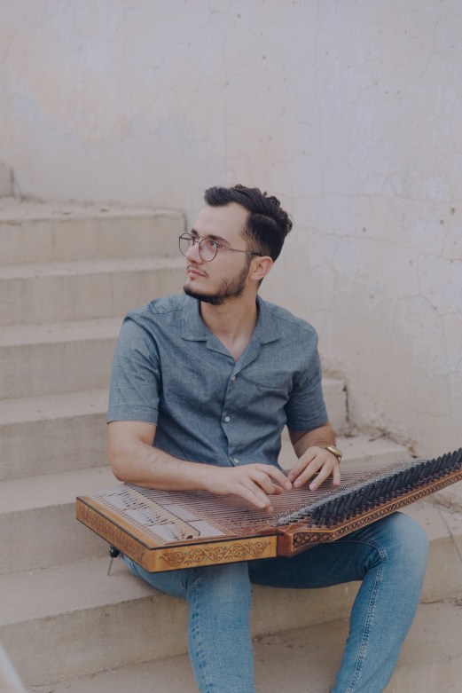 Young man with glasses playing an ornate stringed instrument while sitting on stone steps