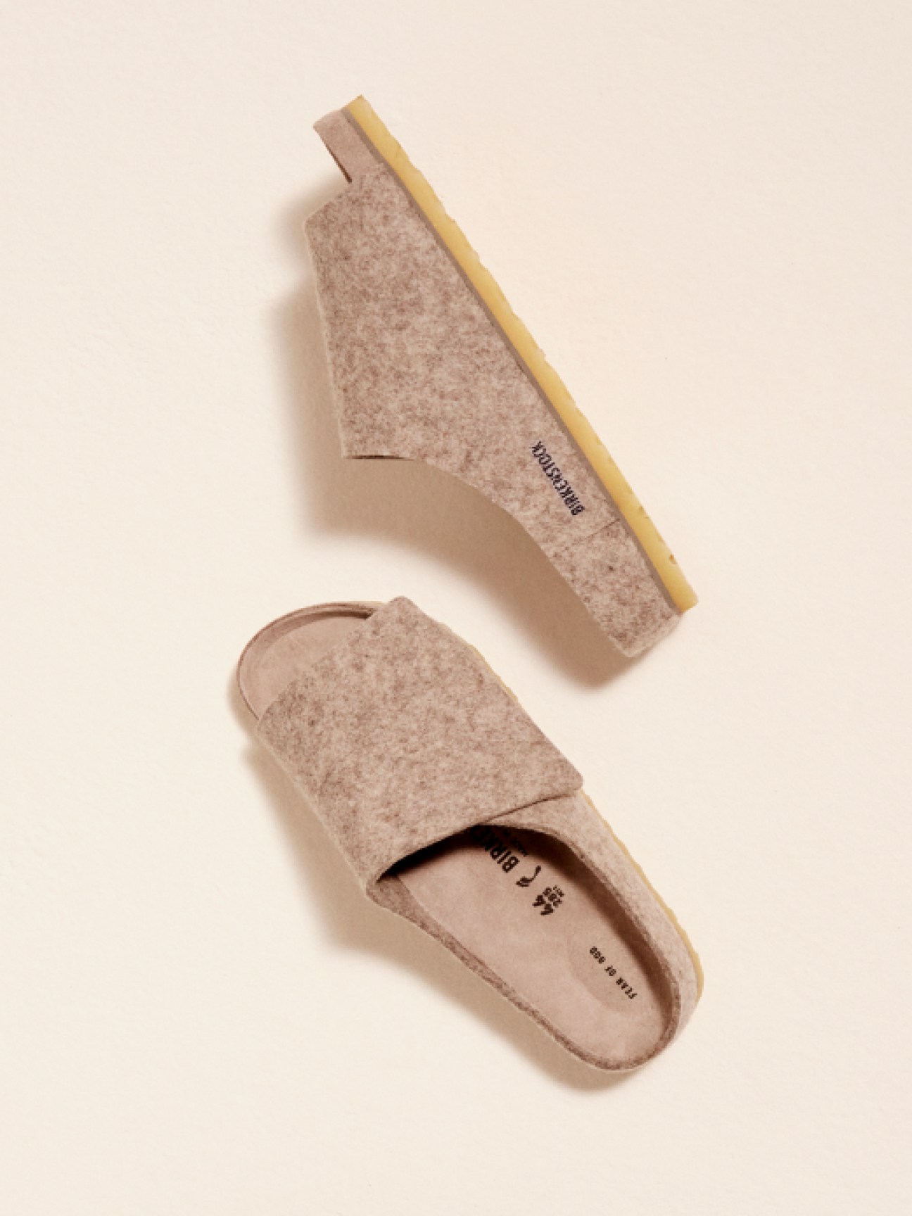 Two beige felt slippers with cork soles photographed against a cream background, one upright and one flat