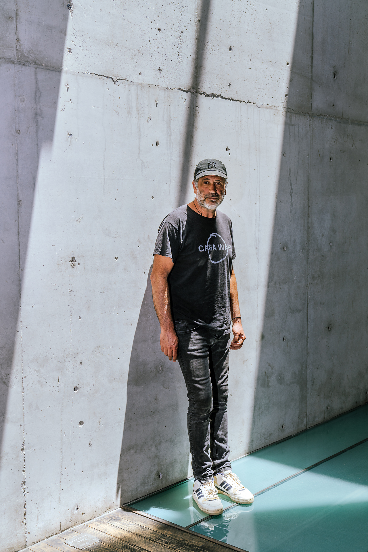 Man in black "Casa Wabi" t-shirt and cap standing against concrete wall with shadow