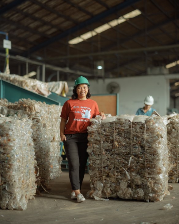 Dian Kurniawati standing among large piles of collected plastic waste at Tridi Oasis recycling facility, demonstrating the scale of plastic waste processing