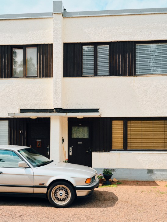 Cream-colored modernist terraced housing with dark wood shutters and a vintage BMW parked outside