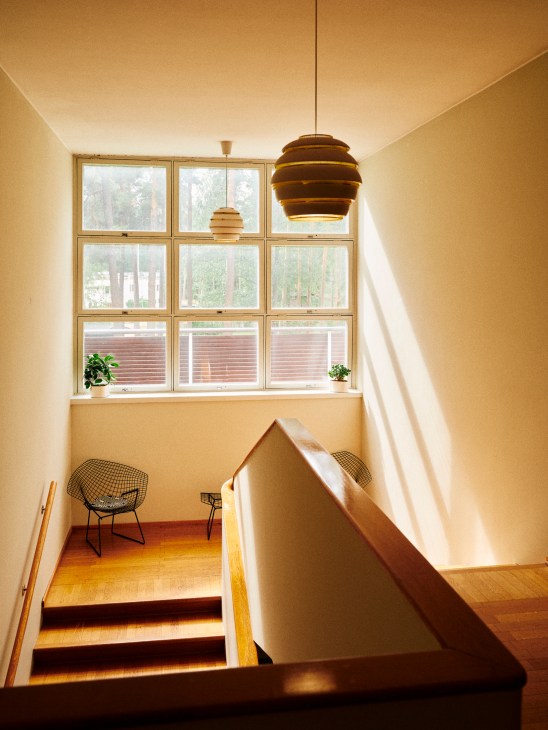Bright staircase landing with modernist pendant light, wire chairs, and large grid windows overlooking forest