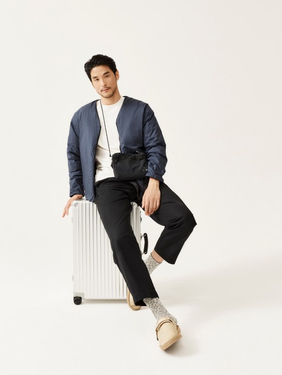 Man in navy jacket and black pants sitting on white suitcase with crossbody bag and sandals
