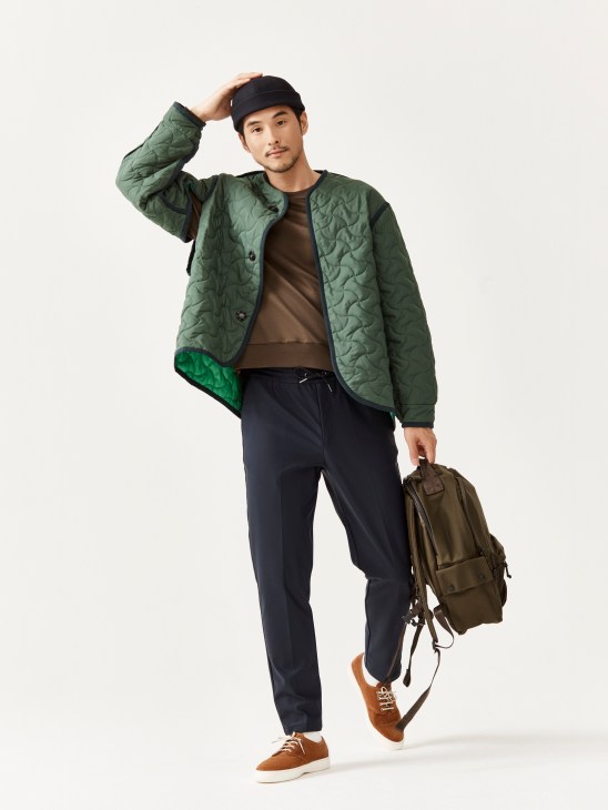 Man in green quilted jacket, brown shirt, navy trousers and beanie poses with olive backpack against white background