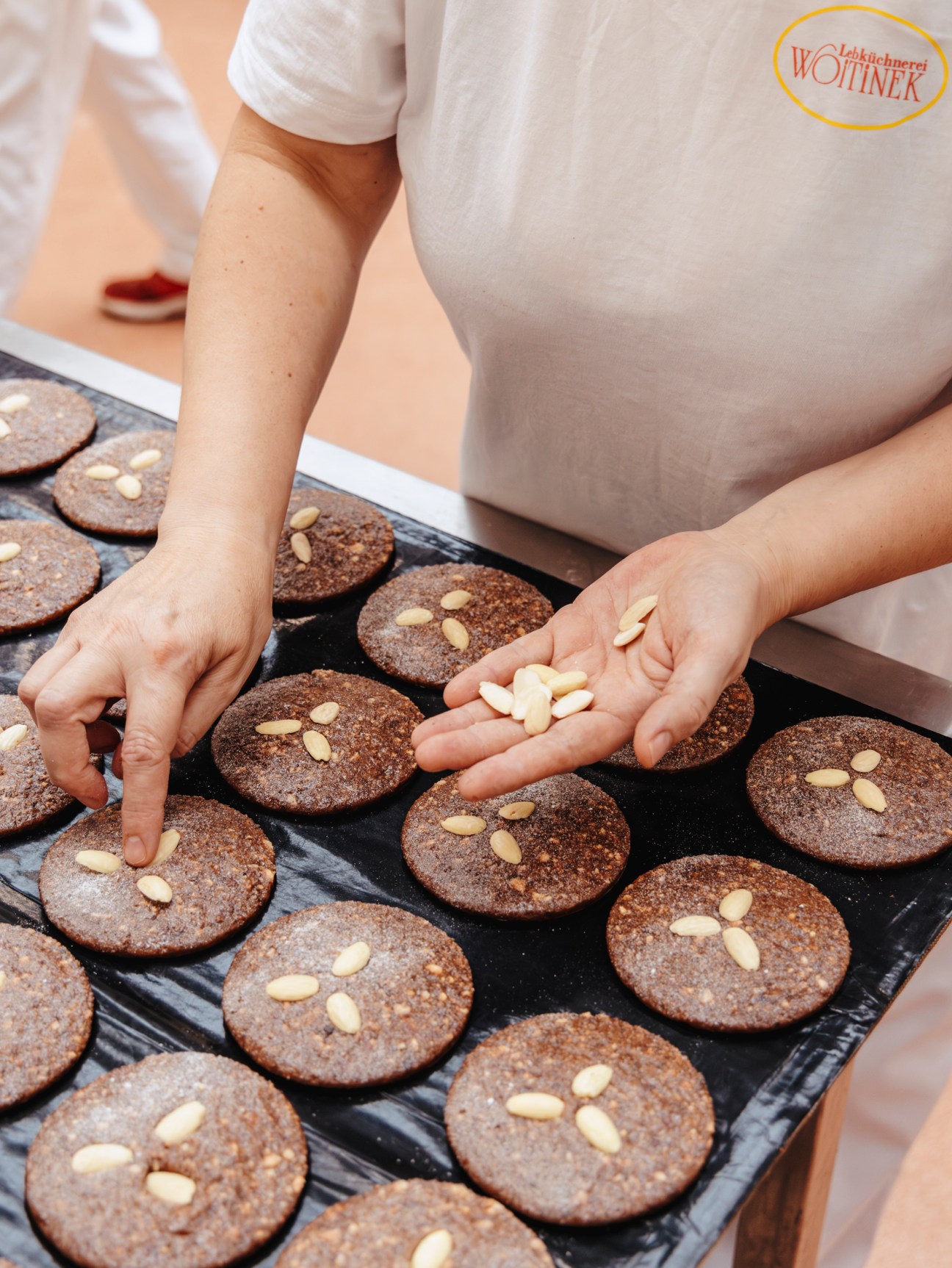 Hands placing sliced almonds on dark chocolate Lebkuchen cookies arranged on black baking trays