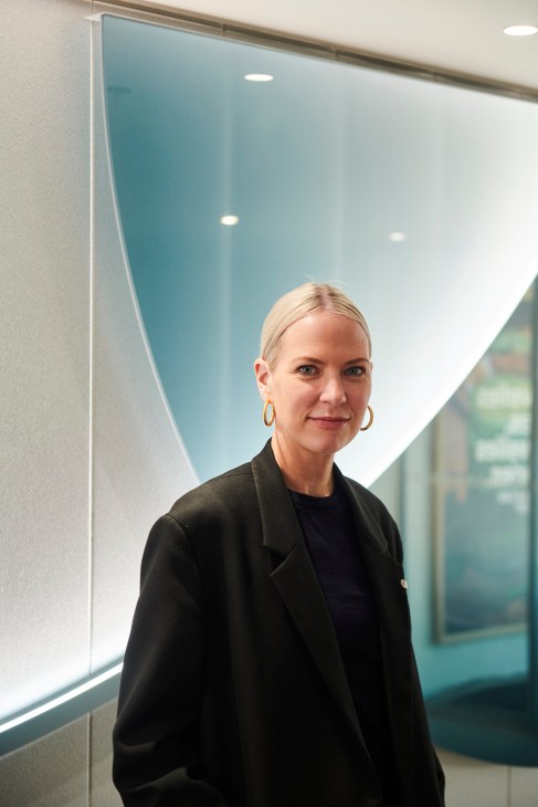 Blonde woman in black blazer and gold hoop earrings standing in modern office with glass walls