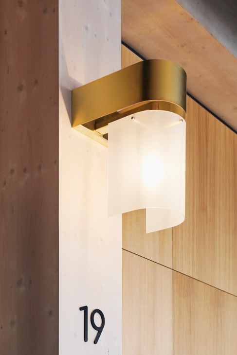 Modern cylindrical brass wall sconce with white lampshade mounted on light wood wall next to number 19