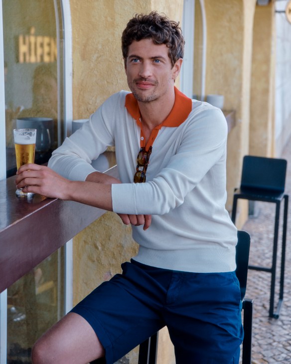 Man in white polo shirt and beige shorts with sunglasses against yellow wall
