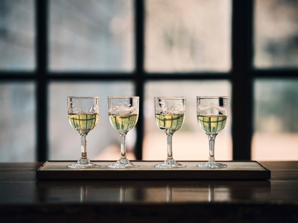 Four etched glass wine glasses with white wine arranged on a wooden tray by a window