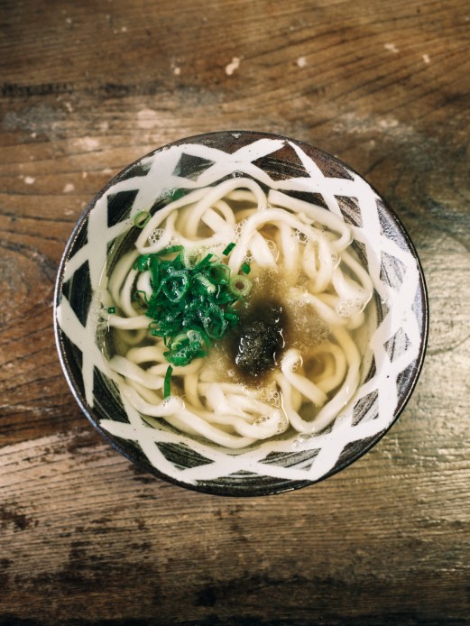 Bowl of udon noodles in clear broth with green seaweed garnish served in decorative ceramic bowl