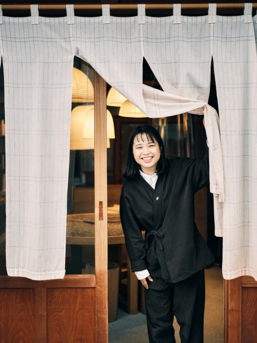 Smiling woman in black traditional Japanese clothing parting white curtains at entrance to traditional building