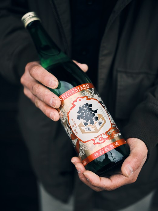 Hands holding a green bottle of Shigemasu sake with Japanese text and red labeling