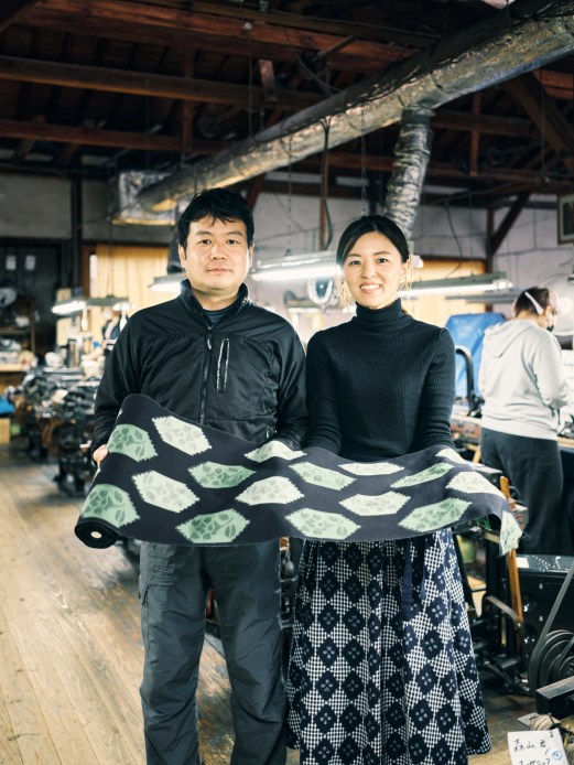 Man and woman holding patterned textile in workshop with exposed ceiling beams