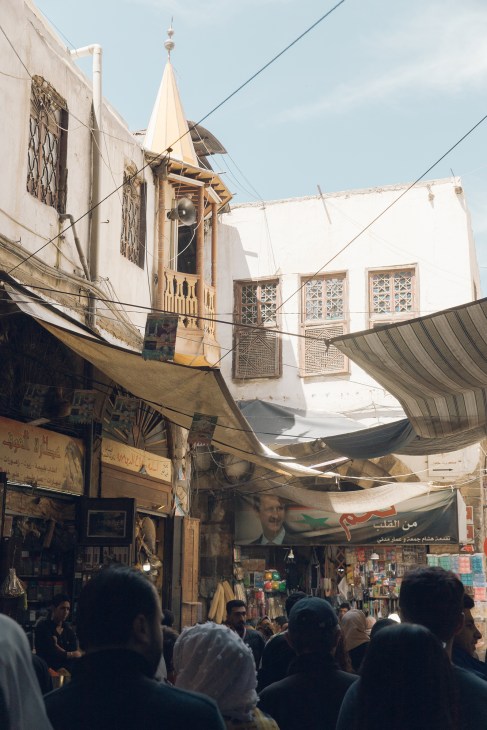 Crowded marketplace in Damascus's Old Town with traditional stone buildings, minaret, and people walking through narrow streets