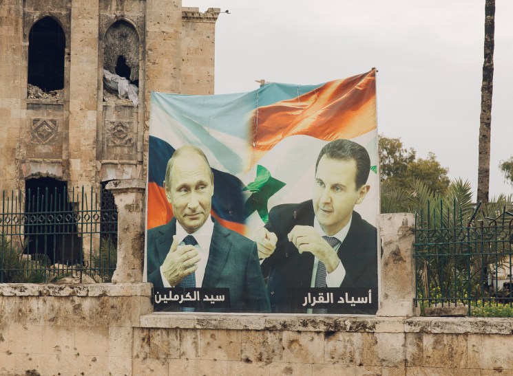 Propaganda posters showing two men in suits displayed on a damaged stone wall with Russian and Syrian flags