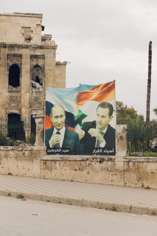 Propaganda posters showing two men in suits displayed on a damaged stone wall with Russian and Syrian flags