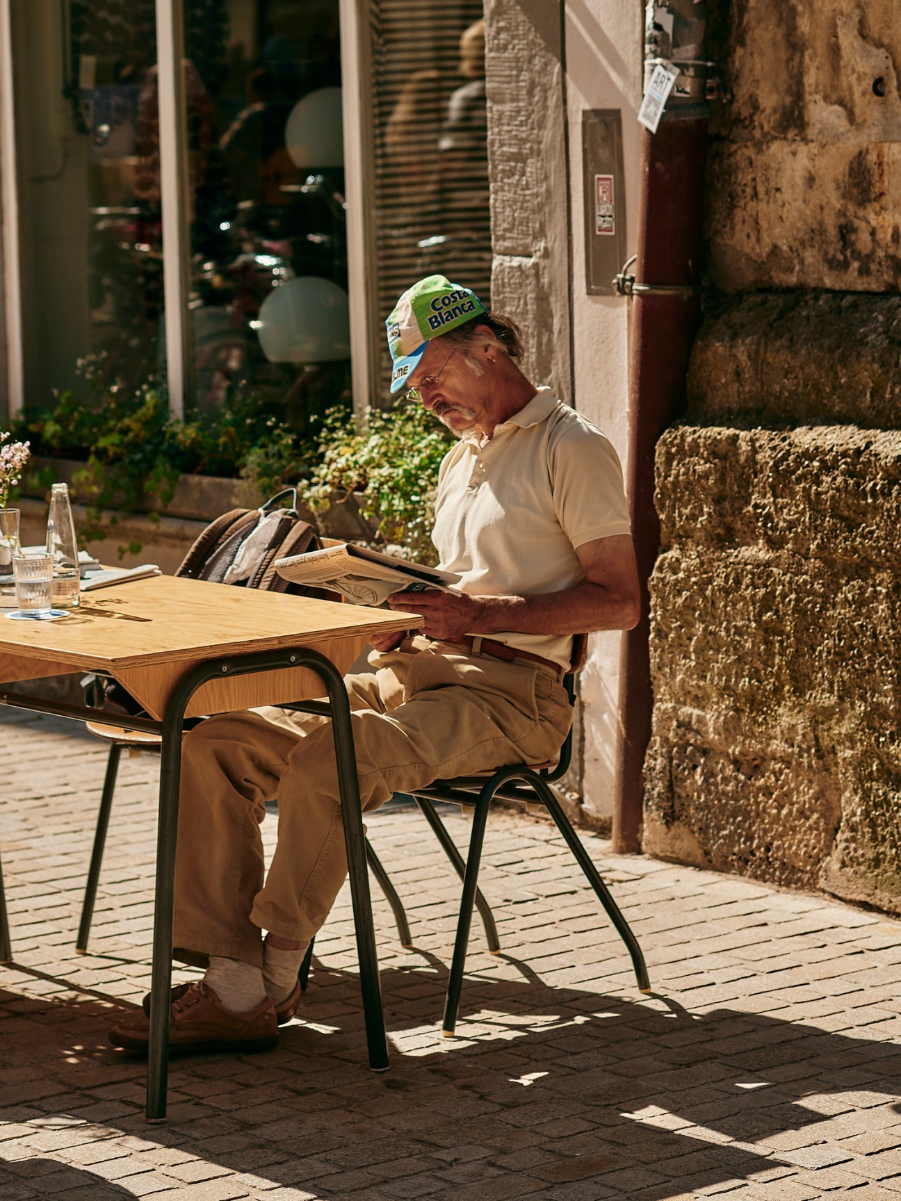 Elderly man in cap reading newspaper at outdoor cafe table in cobblestone square
