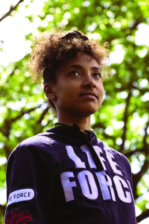 Woman with curly hair wearing a purple hoodie with 'LIFE FORCE' text, photographed outdoors under green foliage