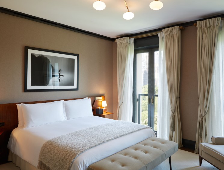 Elegant hotel bedroom with white bedding, neutral tones, large windows with curtains, and city view