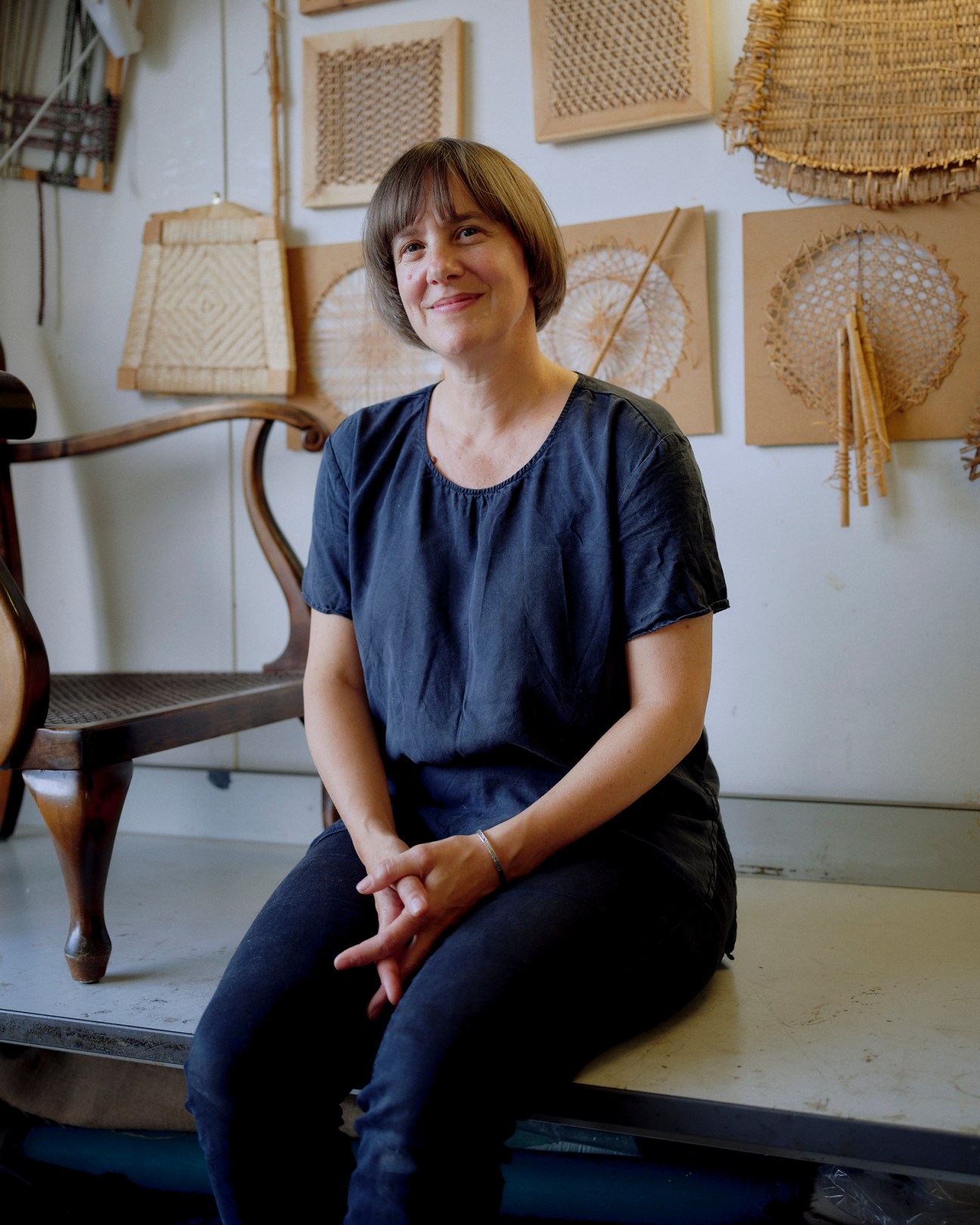 Rachael South sits in her upholstery workshop surrounded by woven chair seating samples displayed on the wall behind her.