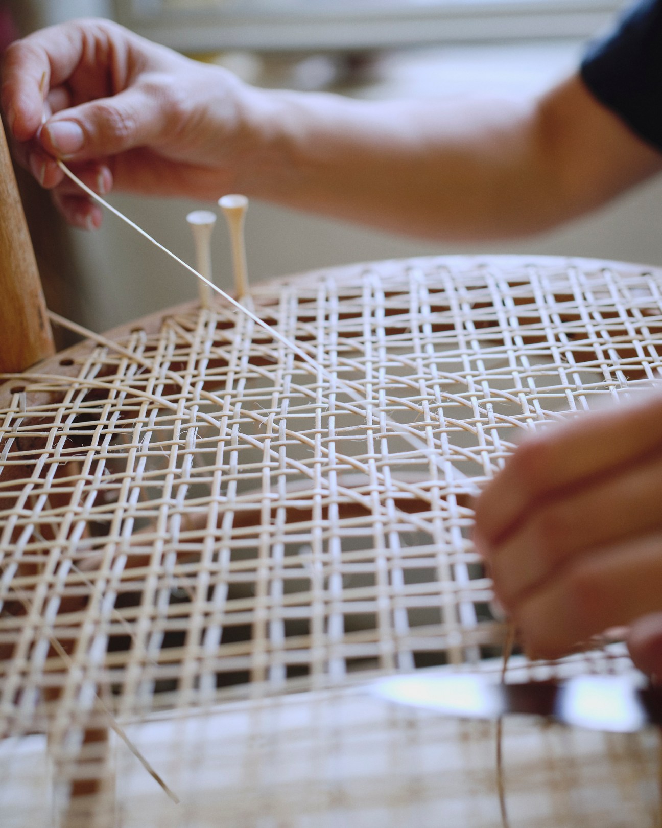 Hands demonstrating cane weaving technique on a woven chair seat pattern.