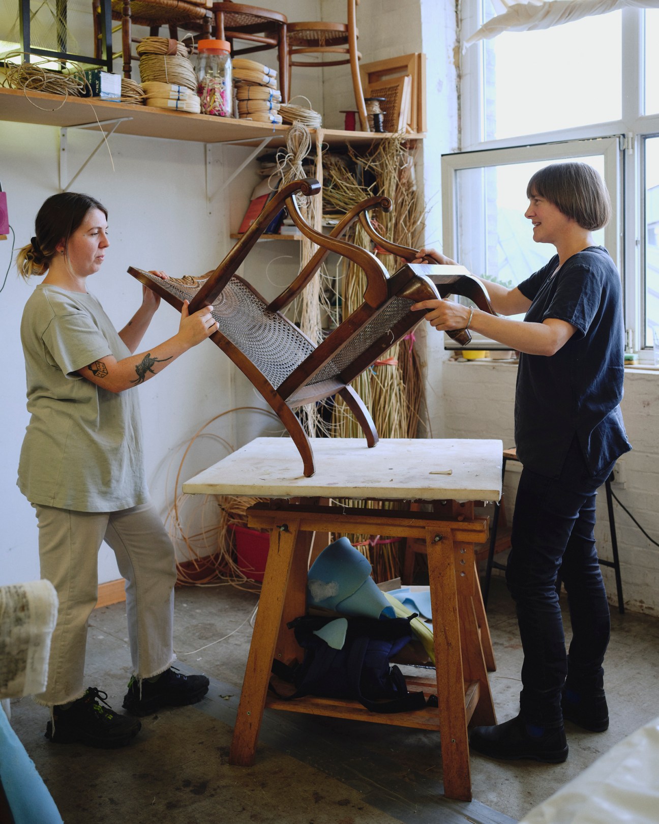 Two women demonstrate chair caning techniques in a craft workshop, working on a wooden chair frame at a work table.