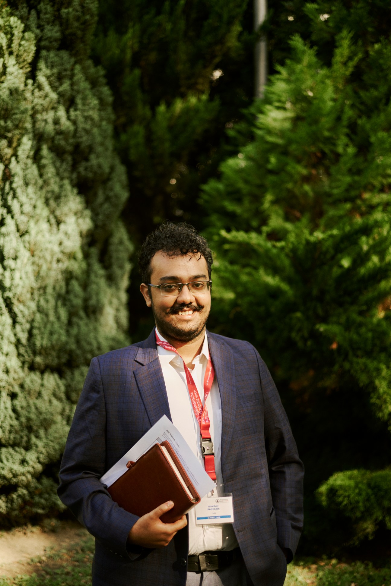 Anubhav Banerjee, an Indian student, standing outdoors with glasses, suit jacket, and holding papers