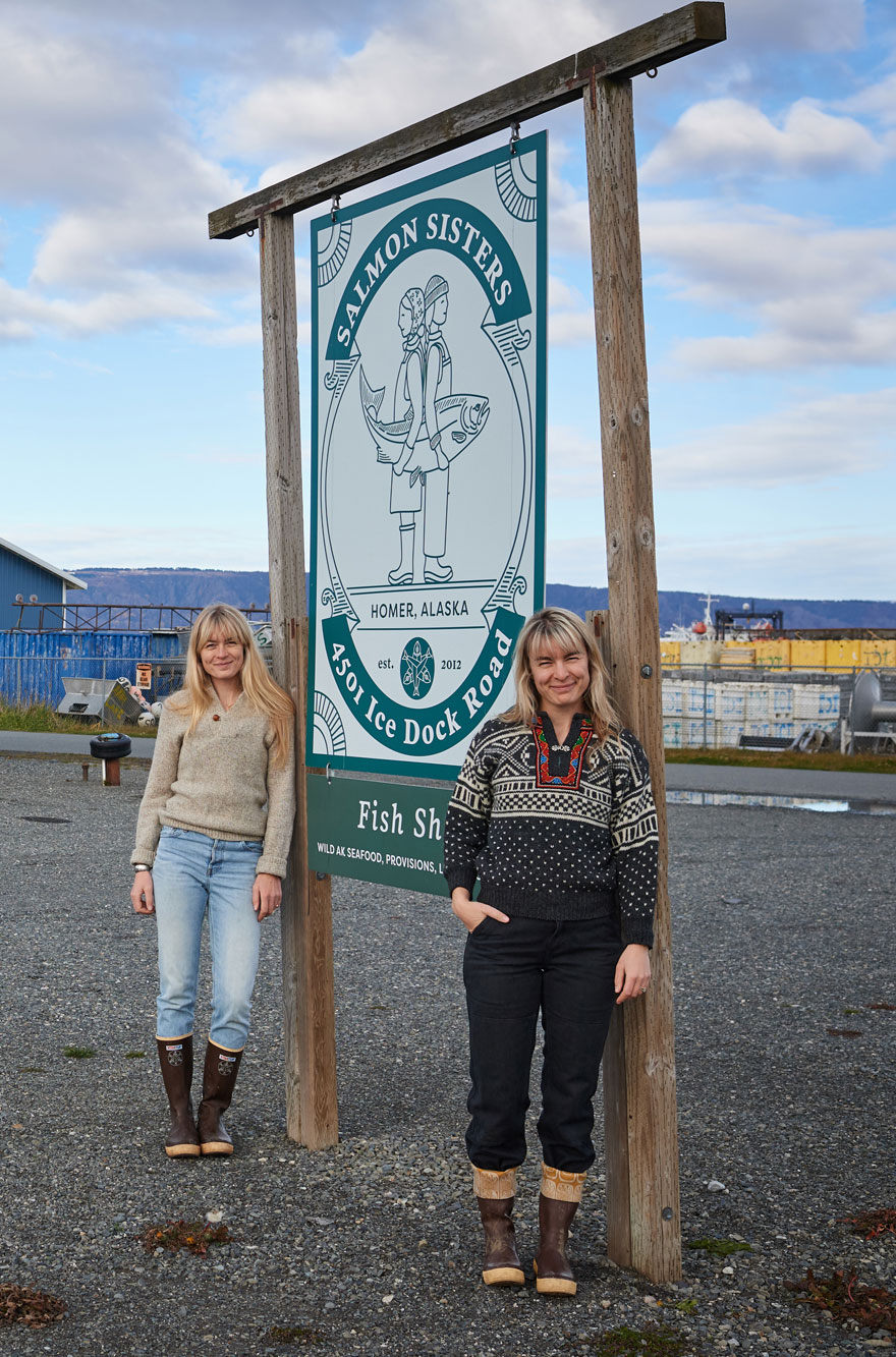Two women in winter clothing standing beside a wooden sign for Salmon Sisters in Homer, Alaska