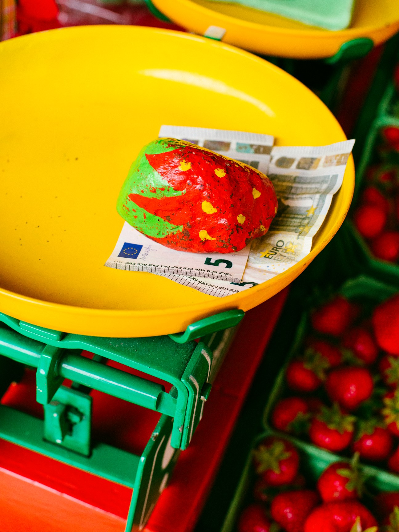 A painted strawberry stone sits on a yellow scale with euro notes, surrounded by fresh strawberries in colorful crates.