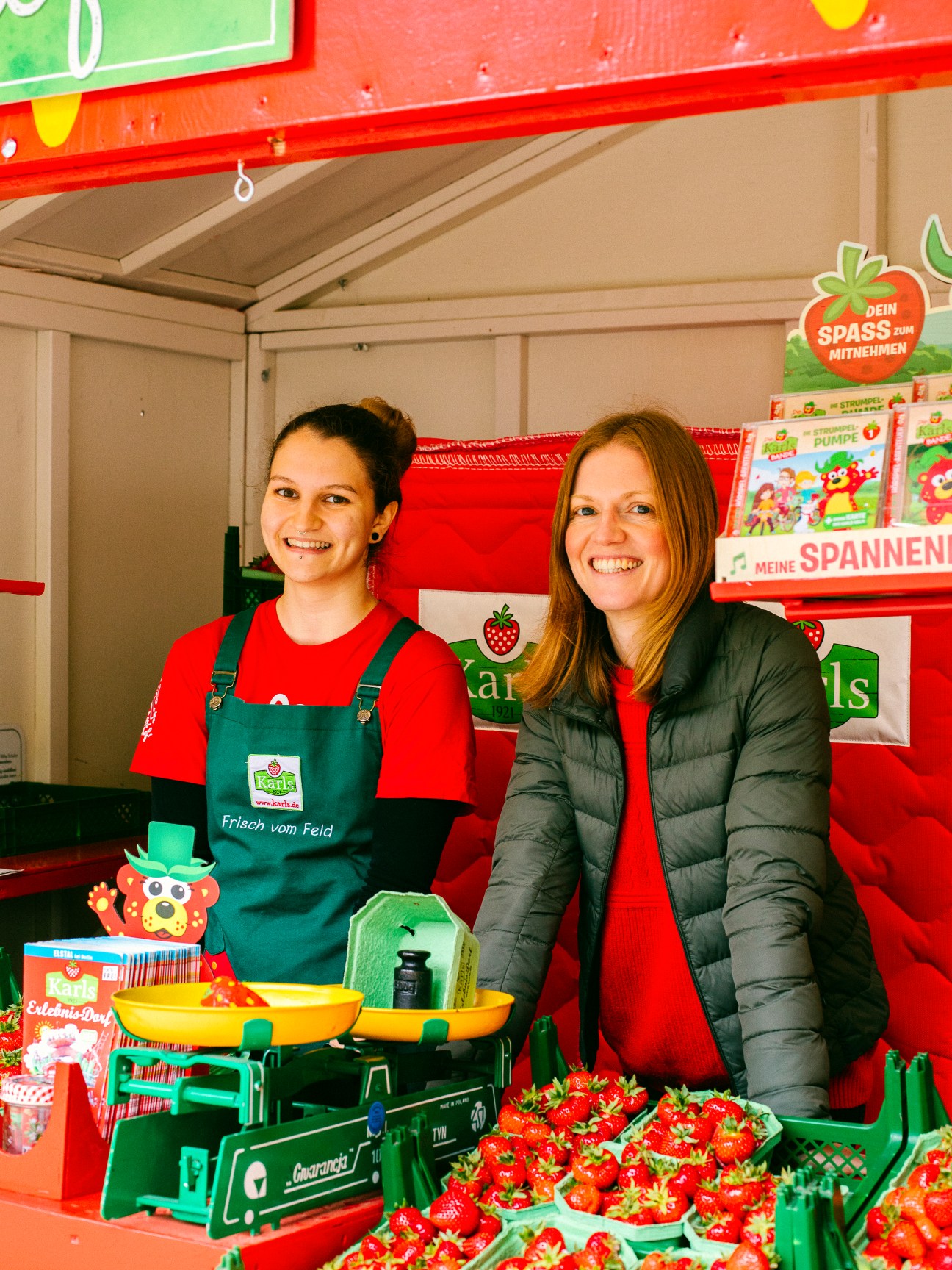 Two women at a Karls strawberry booth in Berlin, with fresh strawberries and branded merchandise displayed.