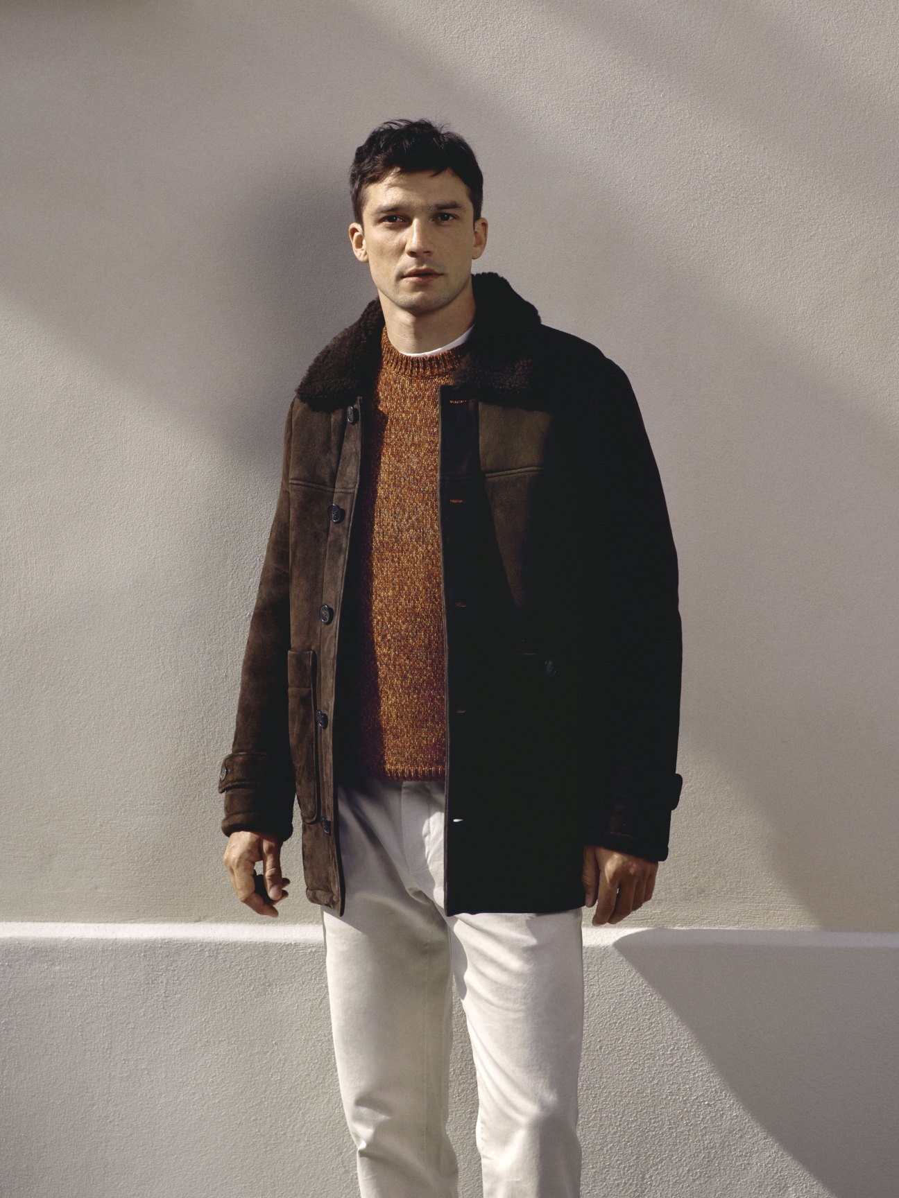 Model in brown leather jacket with fleece collar over orange knit sweater and white trousers