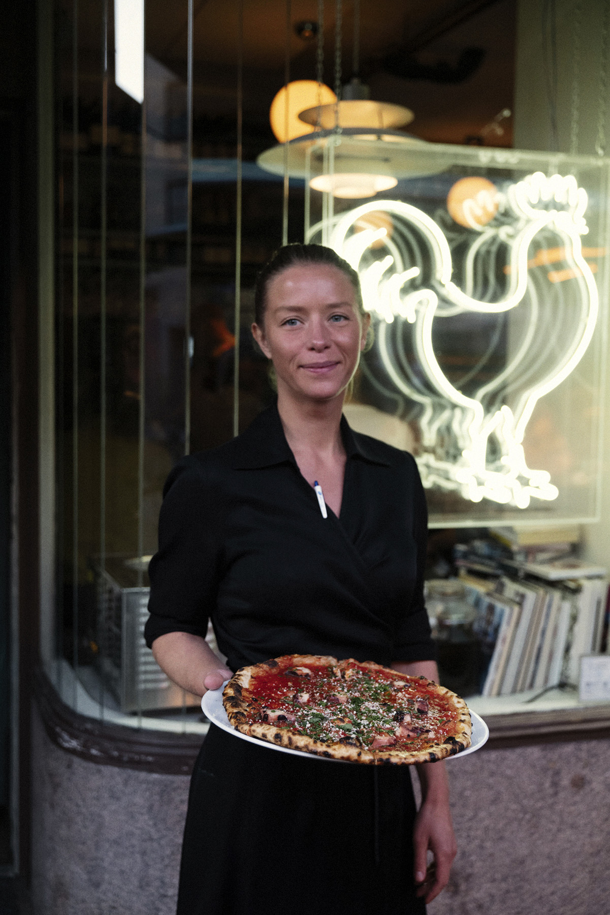 Woman in black holding a pizza outside restaurant with neon sign