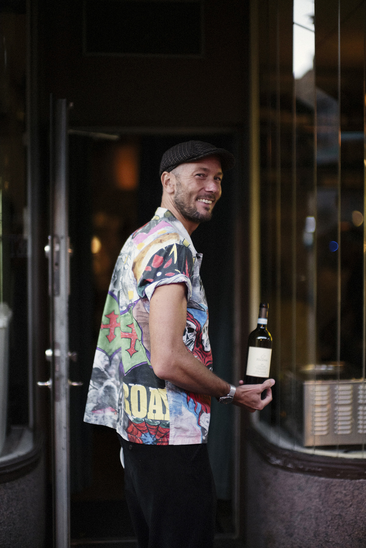 Man in colorful graphic t-shirt and flat cap holding wine bottle outside restaurant entrance