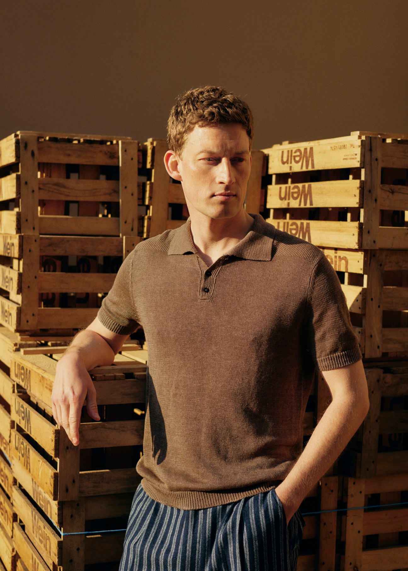 Model wearing brown polo shirt and striped trousers standing against wooden crates