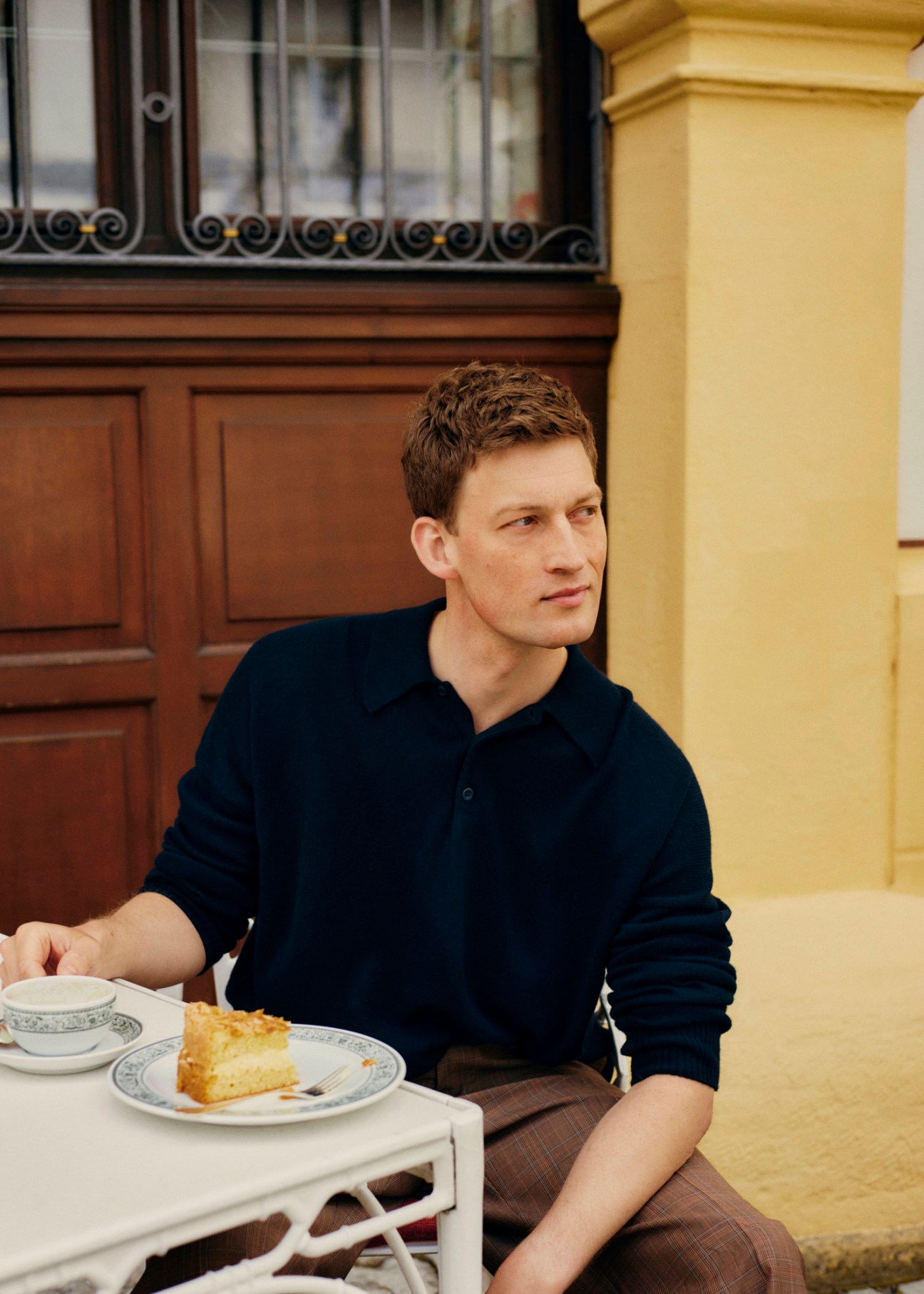 Model in navy knitted polo shirt and brown trousers sitting at cafe table with cake