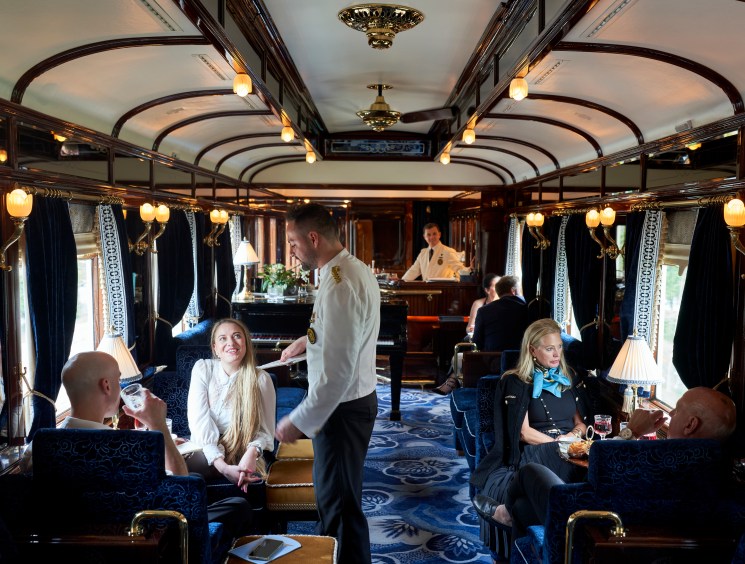 Passengers enjoying drinks in an ornate train bar car with blue upholstery, warm lighting and classic decor