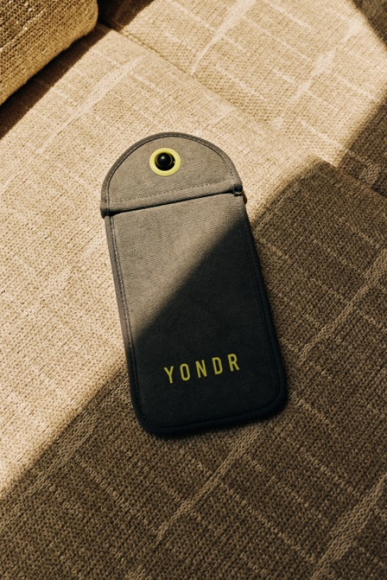 A gray and black Yondr phone pouch with yellow circular lock mechanism lying on a beige textured couch