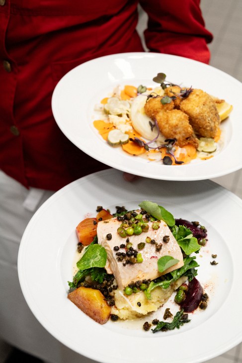 Two white plates of gourmet food - one with breaded items and vegetables, another with fish and greens