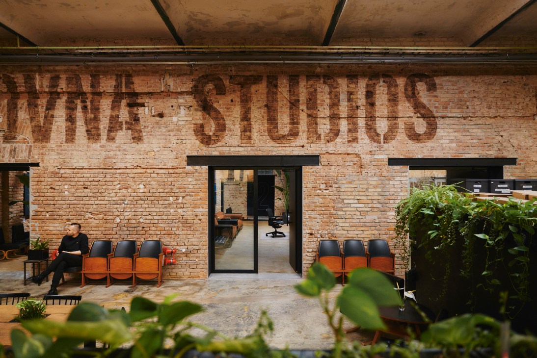 Interior of Kabelovna Studios showing exposed brick wall with faded signage, black-framed glass doors, orange chairs, and a person sitting in the converted cable factory space.