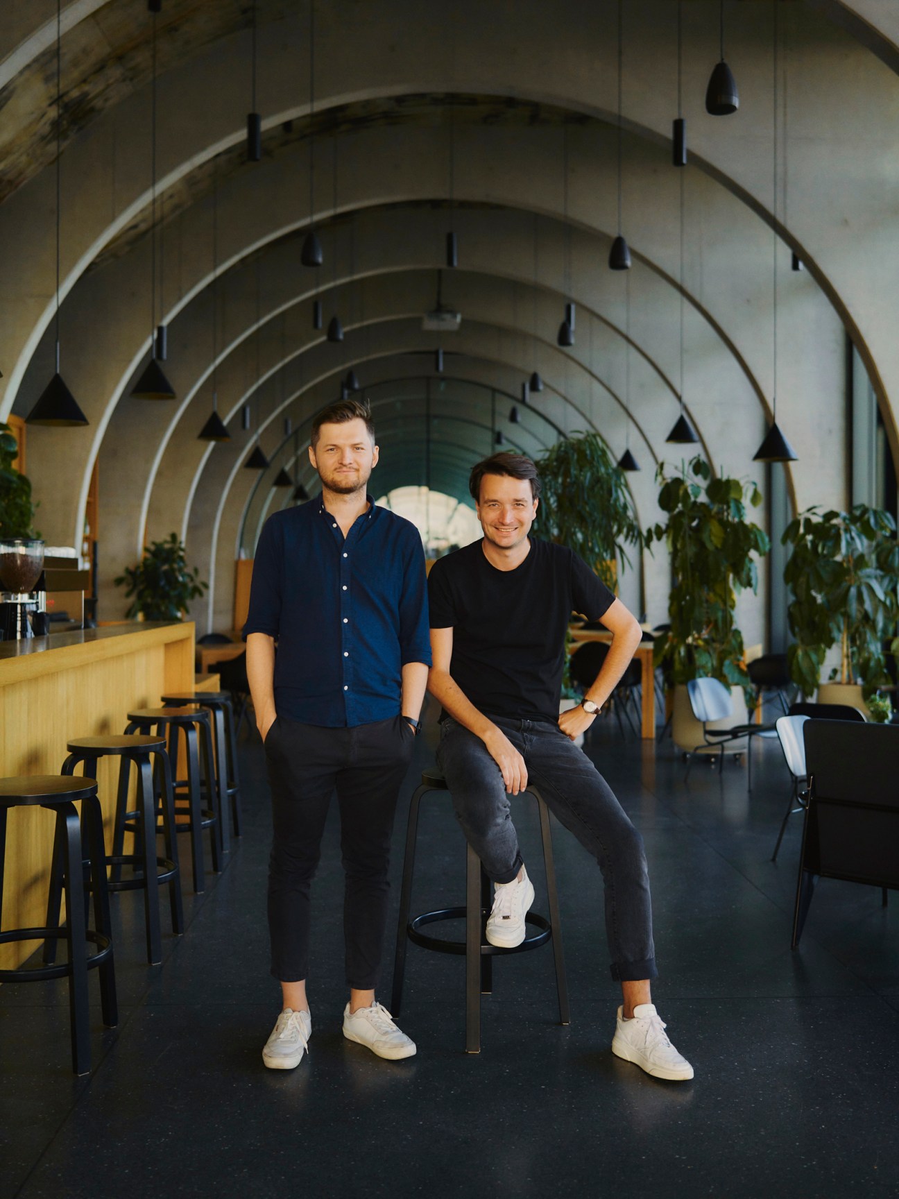 Two men, Ondrej Chybik and Michal Kristof, stand in a modern corridor with curved white ceiling arches and hanging pendant lights, flanked by potted plants.