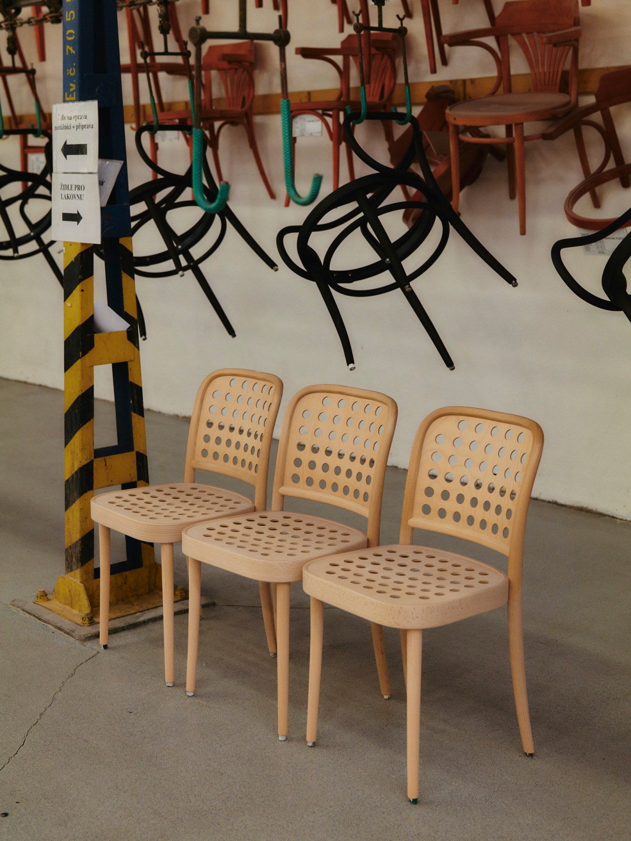Three light wooden chairs with perforated backs and seats arranged in a row, displayed in an industrial space with black sculptural furniture visible in the background.