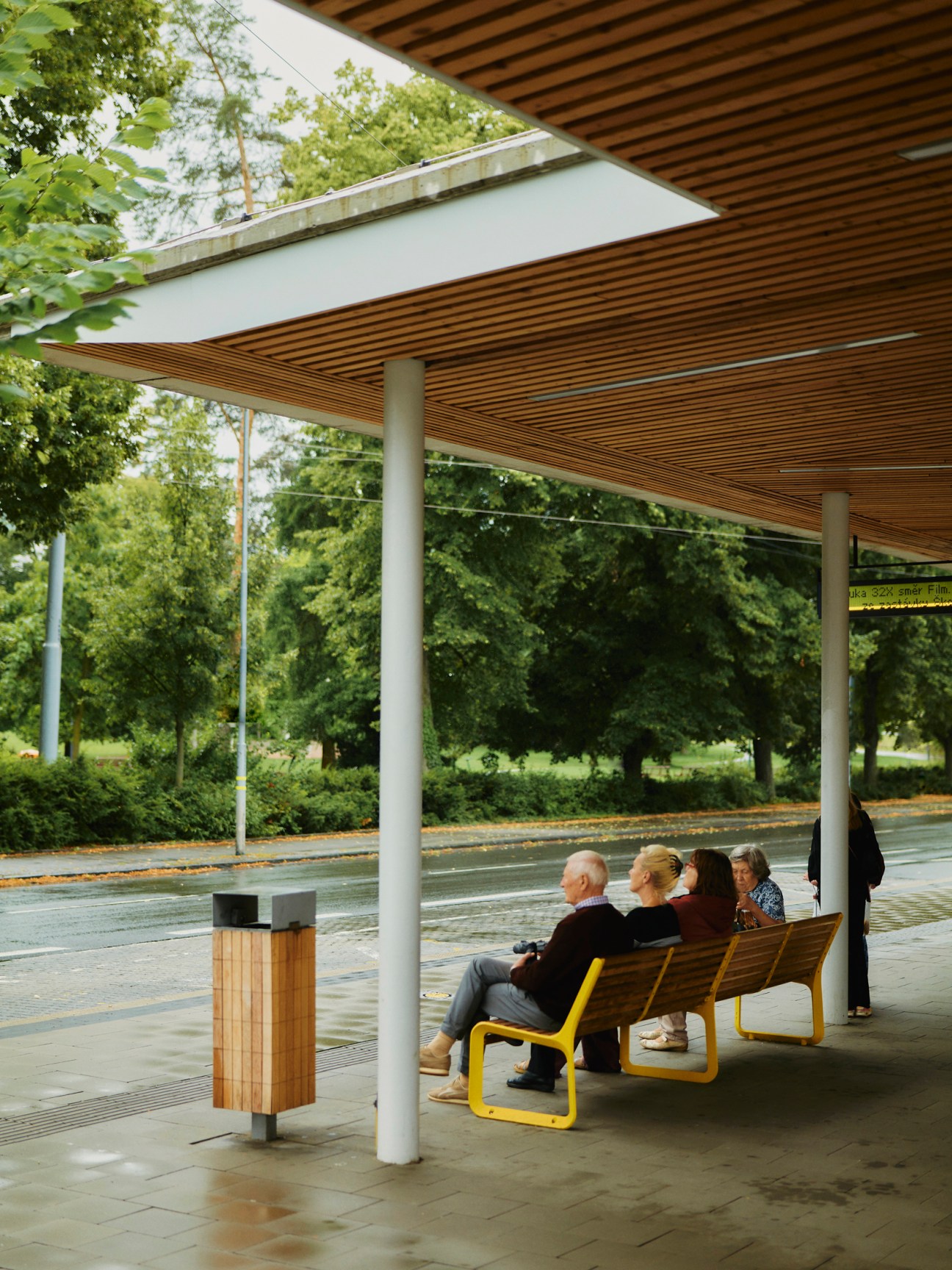 People sitting on modern wooden and yellow metal benches under a contemporary shelter with slatted wooden ceiling