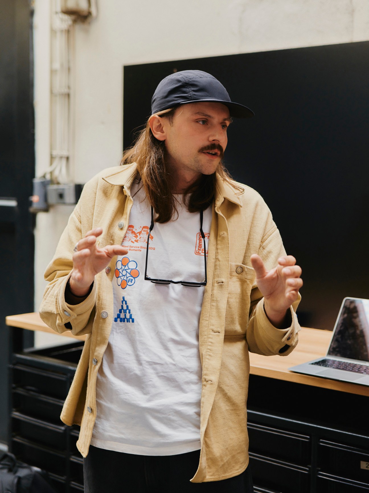 Robin Fiser, with long hair and baseball cap, gesturing while speaking, wearing beige shirt over white t-shirt in modern office