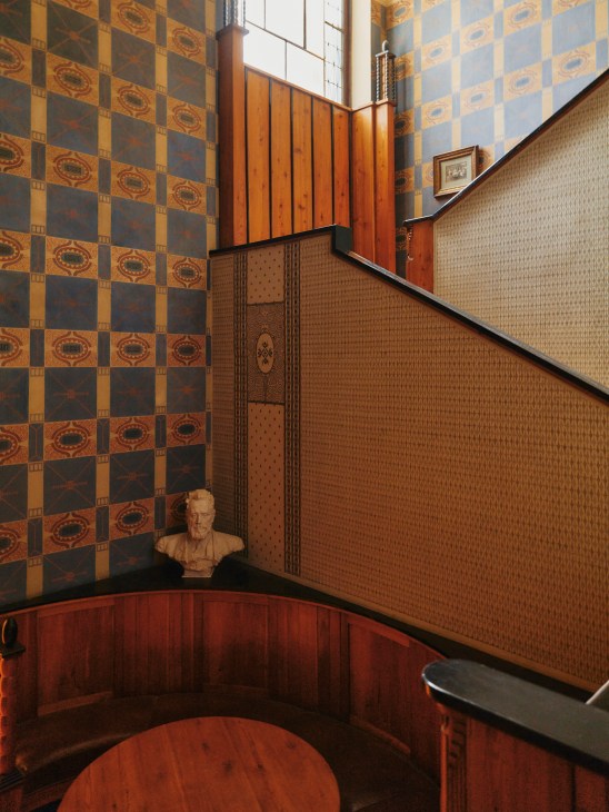 Interior corner of a stairwell featuring a checkerboard patterned wallpaper in gold and dark tones on the upper left wall, wood paneling and textured wallcovering on the right, wooden wainscoting, a classical bust sculpture on a shelf, and wooden architectural details including framed panels above.