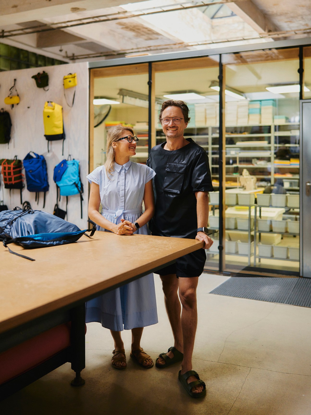 Two people standing at a wooden counter in an outdoor gear shop, with backpacks and climbing equipment displayed on the wall behind them, and glass doors to an interior room visible in the background.
